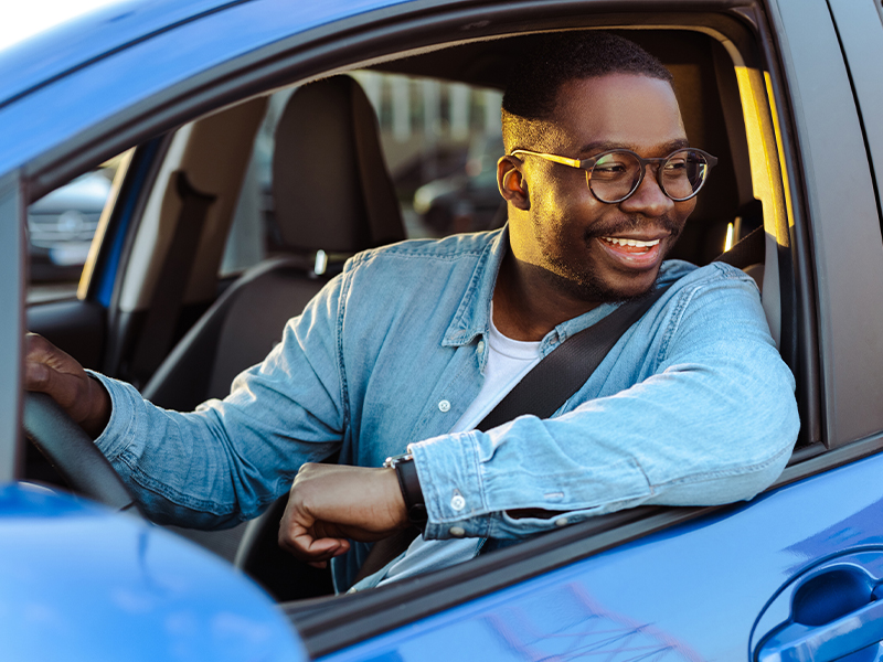 A smiling man in a denim shirt sits in the driver's seat of a blue car, enjoying a sunny day while driving.