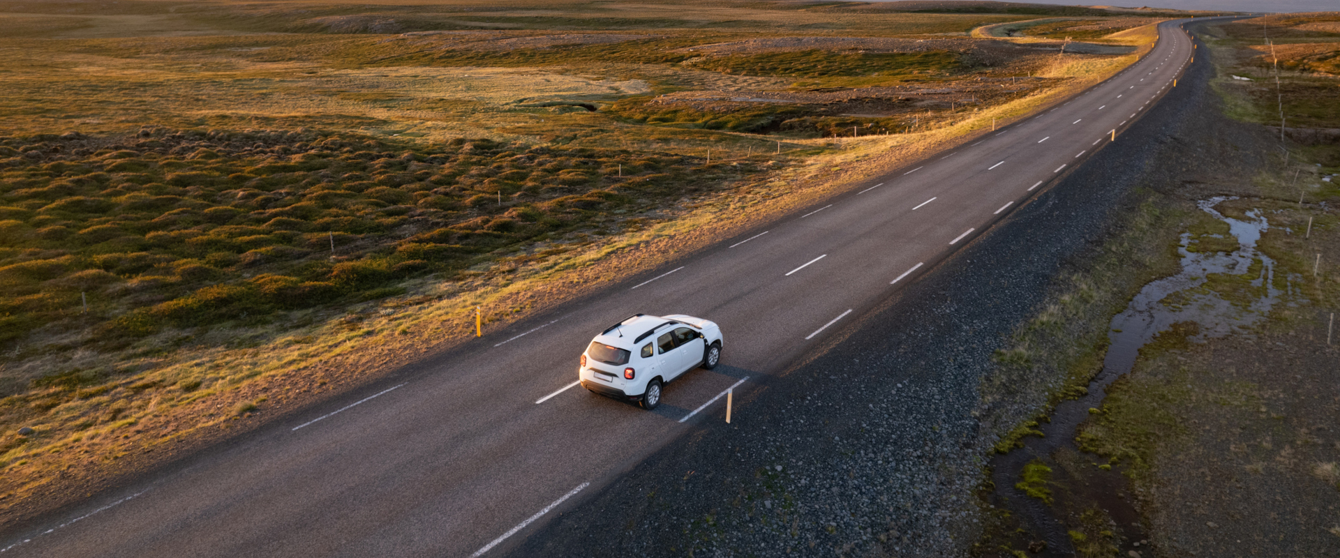 A lone white SUV travels on a long, empty road through a vast, serene landscape at sunset. The road curves gently, bordered by fields and distant mountains.