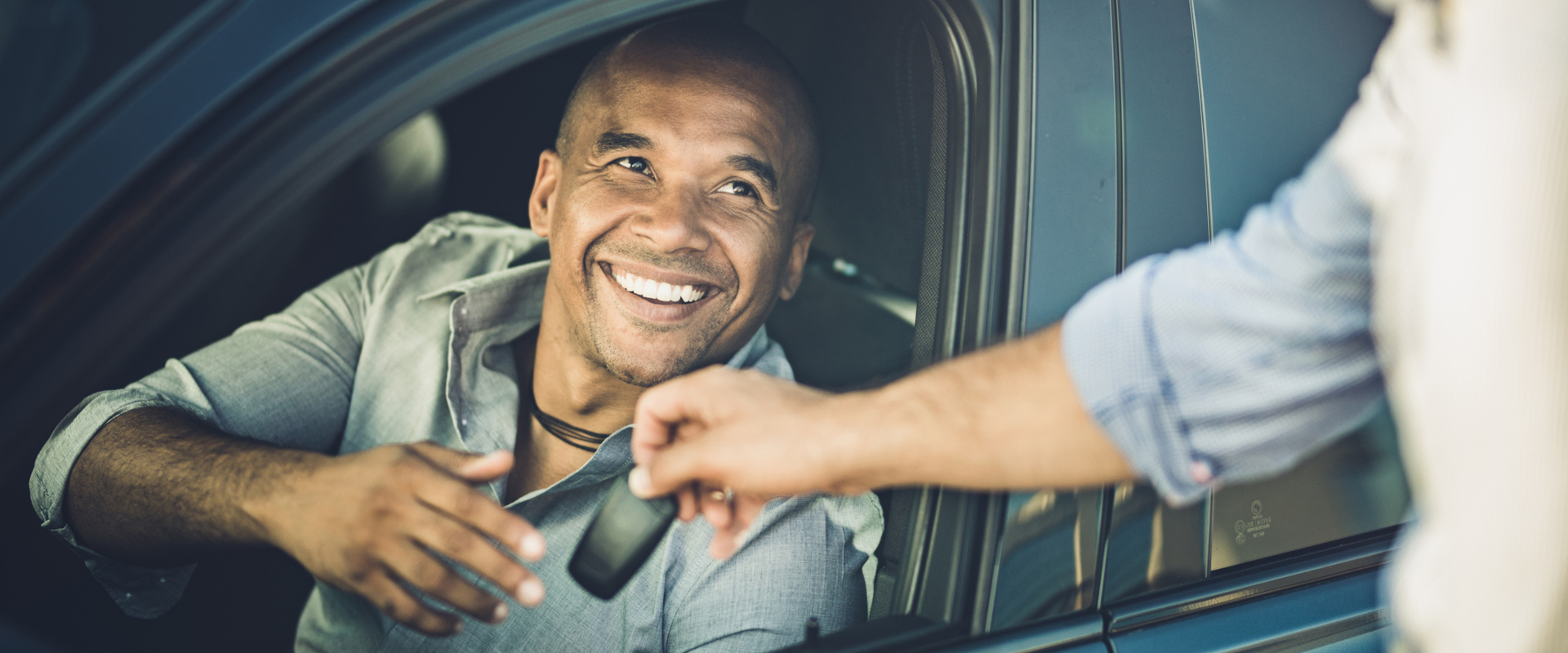 A smiling man in a car reaches for keys handed by another person. The scene conveys friendliness and trust, with a warm, cheerful atmosphere.