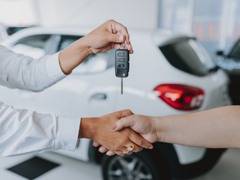 Close-up of a person handing over car keys to another, set in a modern car dealership. The scene conveys a sense of trust and transaction.