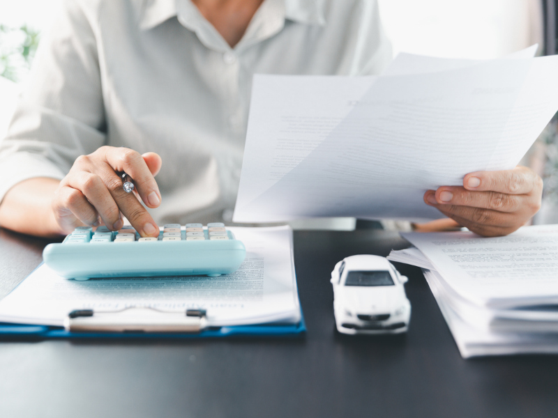 A person reviews documents with one hand while using a blue calculator with the other. A toy car and papers are on the desk, suggesting financial planning.